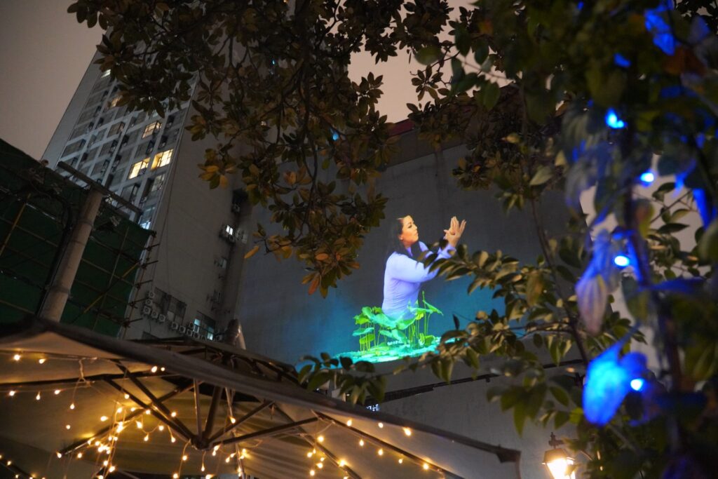 Leaves of a tree are show in the foreground. A projected image of performer Alice Hu is in the background on a wall. Her hands shaped into the form of a lotus.